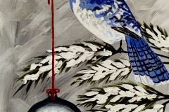 A bluejay sitting on a snowy tree branch holding the string of a Christmas Ornanment in its beak.