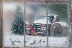 A painting of a snow covered old car parked beside a Christmas tree seen through a window.
